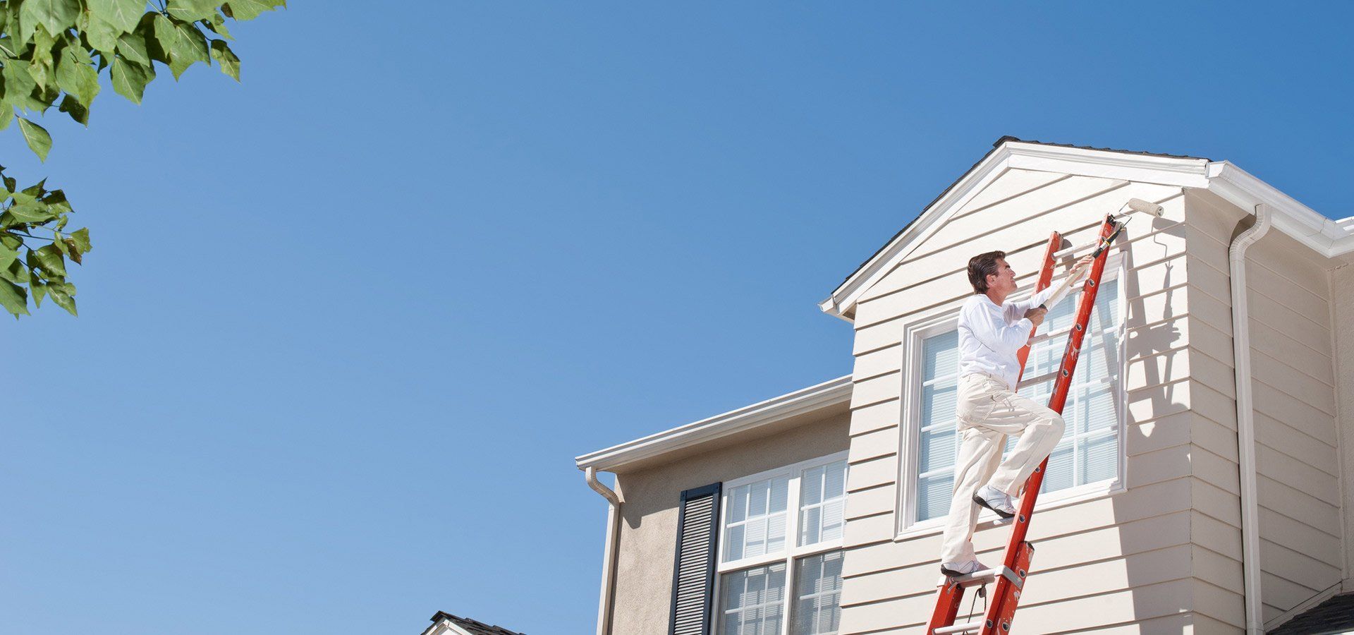 A painter on a ladder paints a house's exterior on a sunny day.