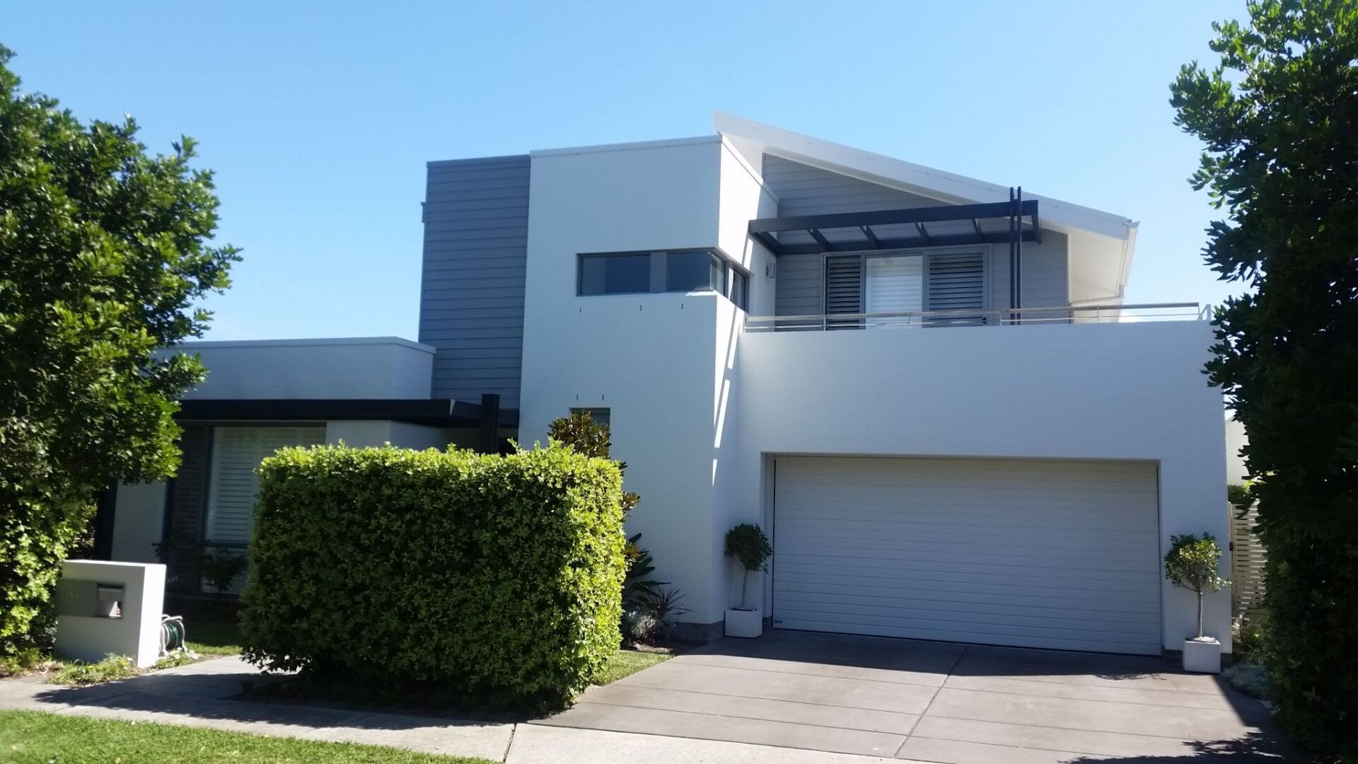 Modern white two-story house with a garage. Light blue sky with surrounding greenery.