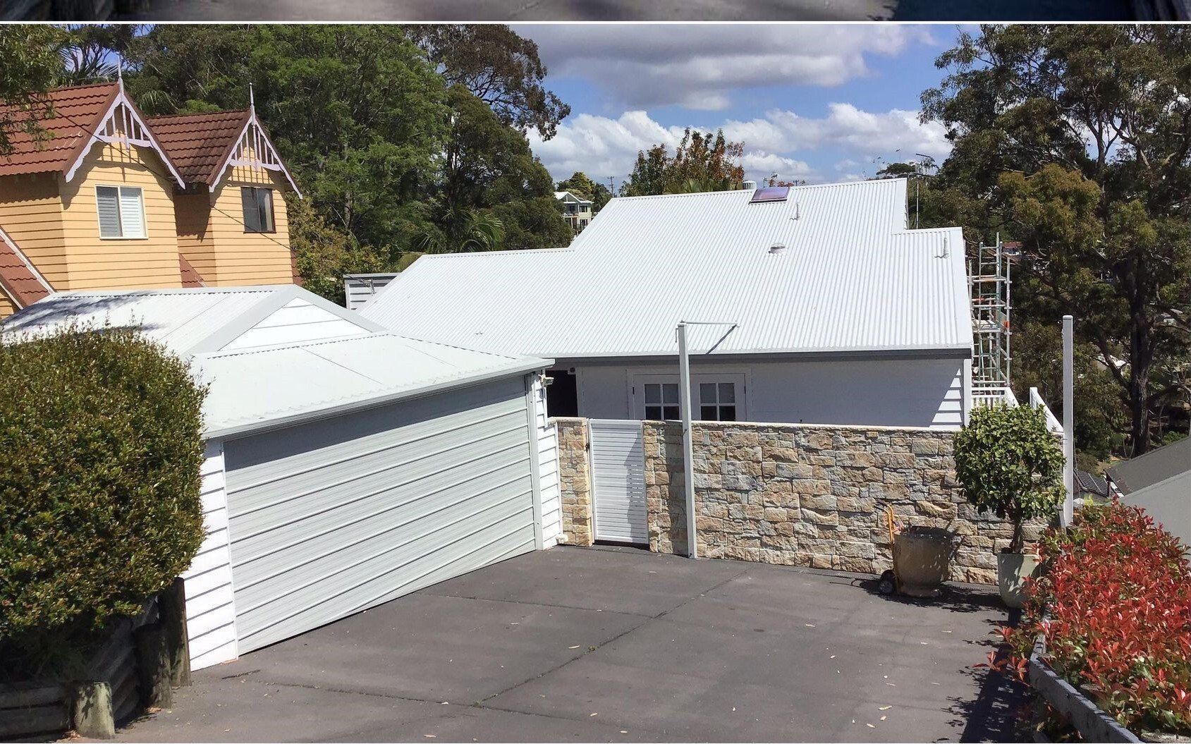 White house with corrugated metal roof and garage, stone wall, asphalt driveway.