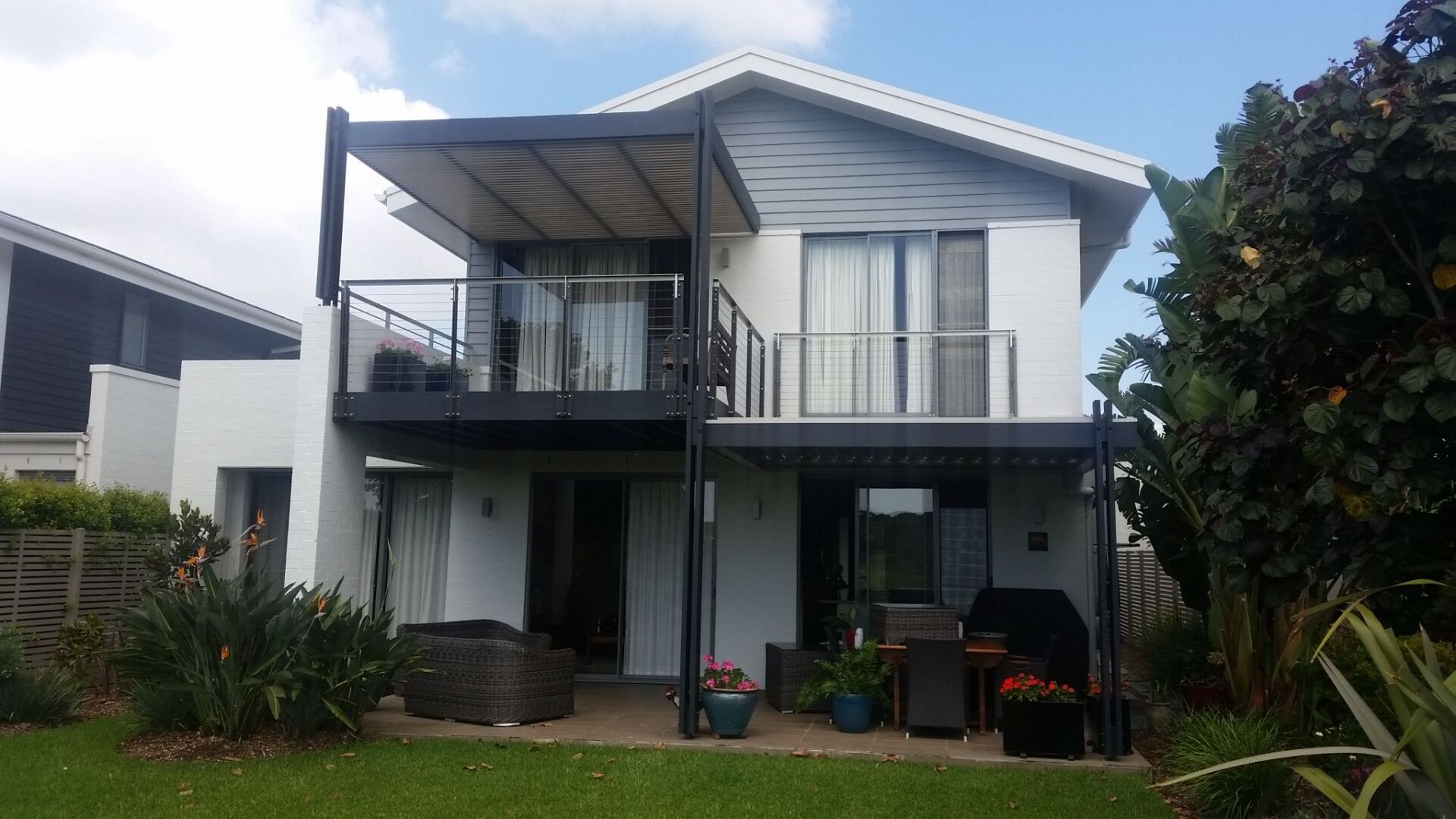 Two-story white house with black balcony and pergola, green lawn and plants.