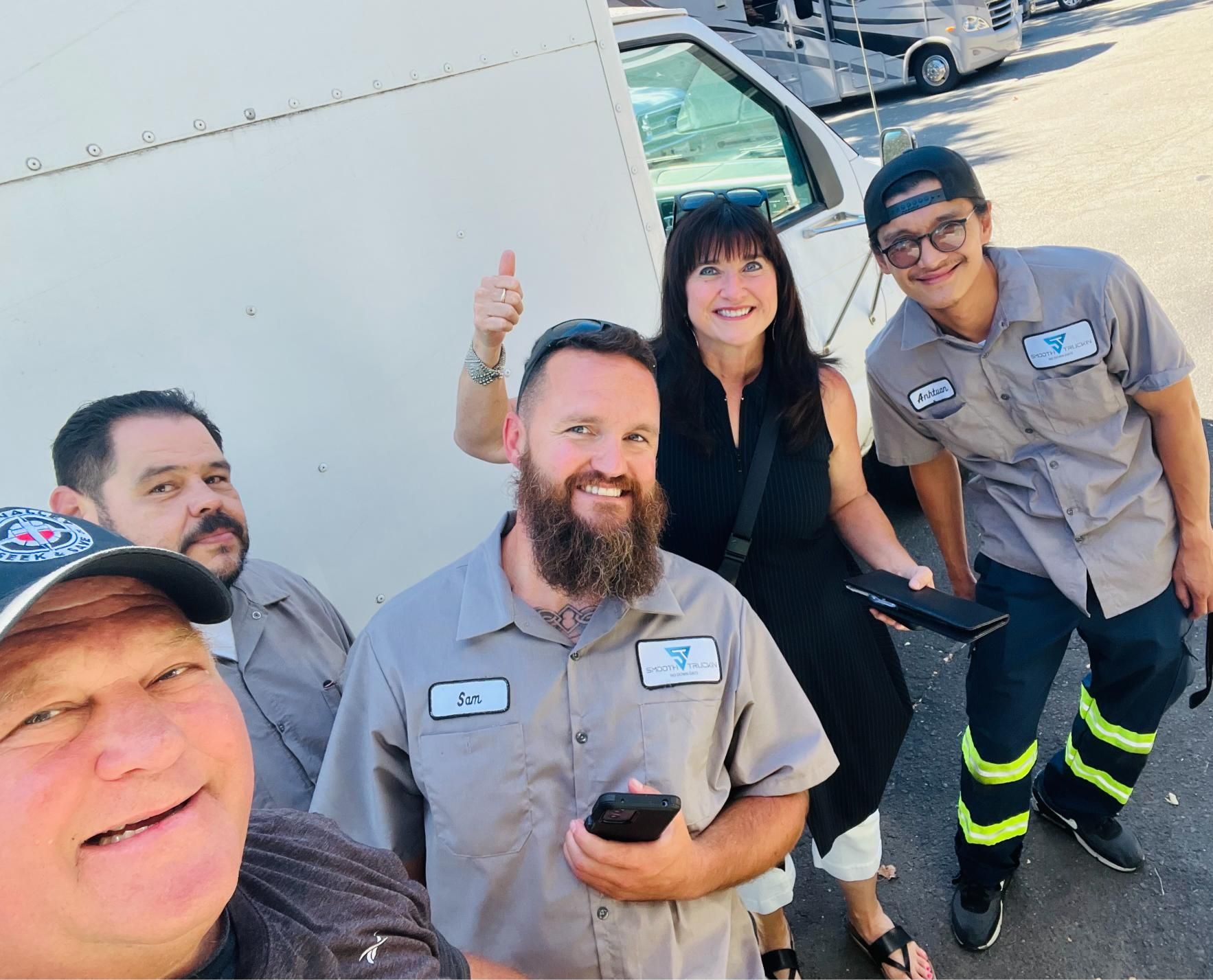 Group of people posing with a vehicle. Smiling, some giving thumbs up. Outdoors, sunny.