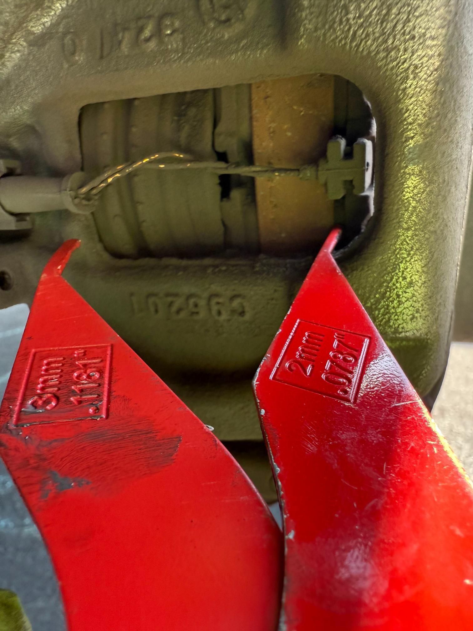 Two red shims measuring 3mm and 2mm, are next to a brake pad, suggesting brake wear measurement.