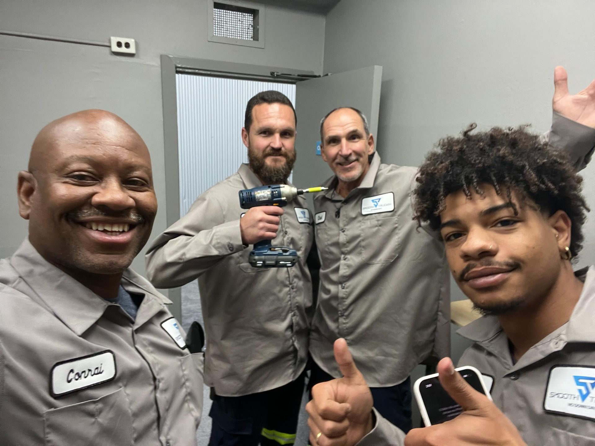 Four men in gray work shirts smiling, one holding a drill. Indoor setting.
