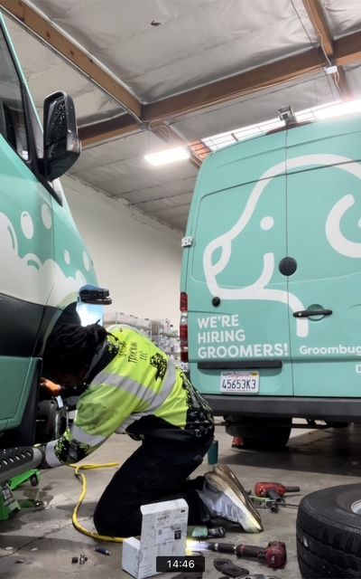 Person kneeling under a turquoise Groombug fleet van, working on the front tire.  Wearing neon yellow vest, in garage.