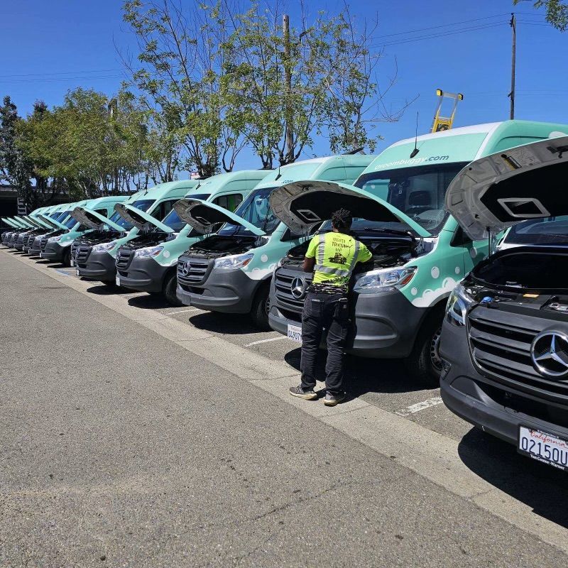 Mechanic inspecting van engines in a row. Vans are green and grey. Sunny outdoor parking lot.