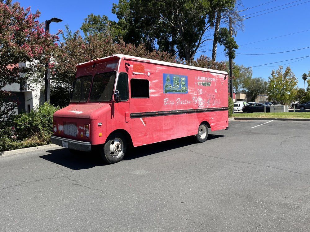 Red fleet truck in a parking lot on a sunny day; blue sign, trees, and cars in background.