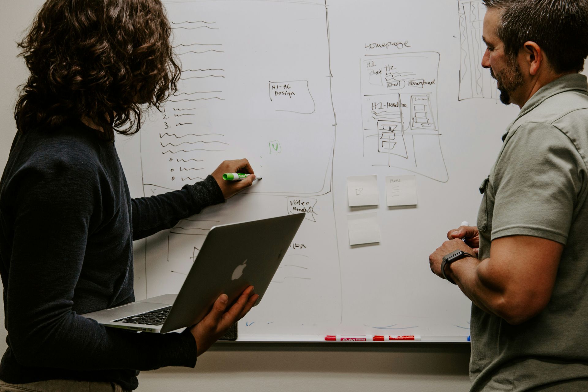 Two people working at a whiteboard with one of the people writing and holding a laptop.