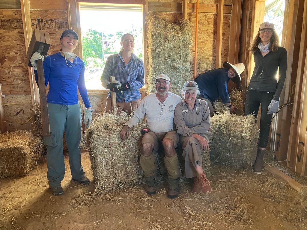 Five people smile while working on a straw-bale building interior, surrounded by loose straw and wooden framing.