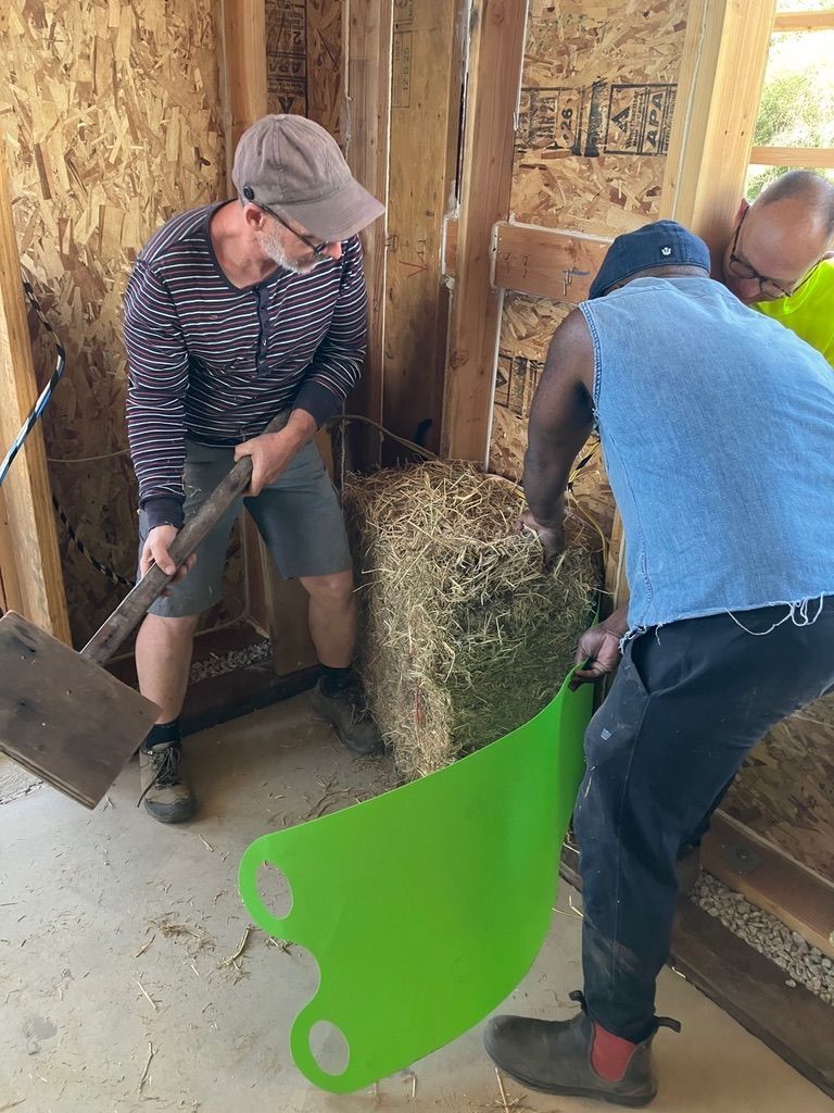 Two people working on a straw bale wall, with one using a spade tool and the other positioning a green guard plate.