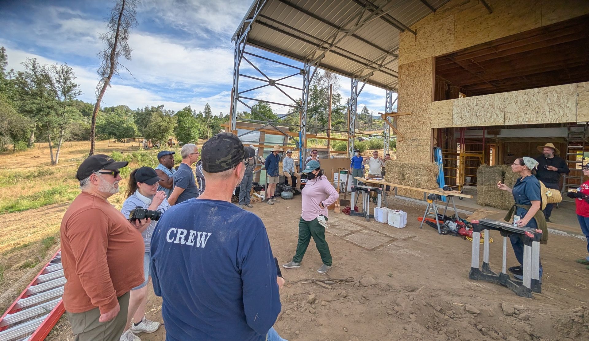 A group of people listens to a speaker at an outdoor construction site with an unfinished wooden structure.