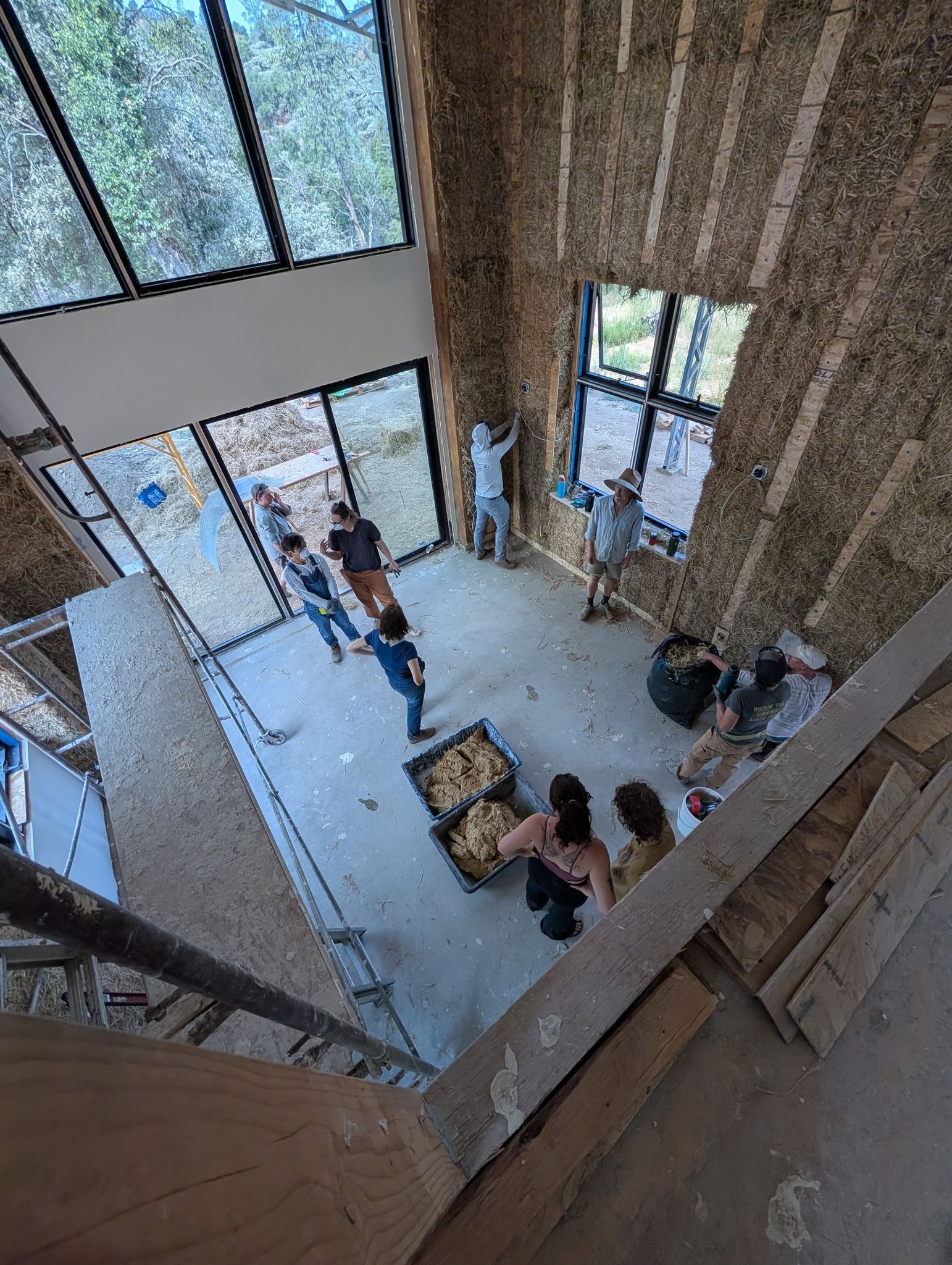 A group of people working inside a high-ceilinged room with exposed straw bale walls and large windows.
