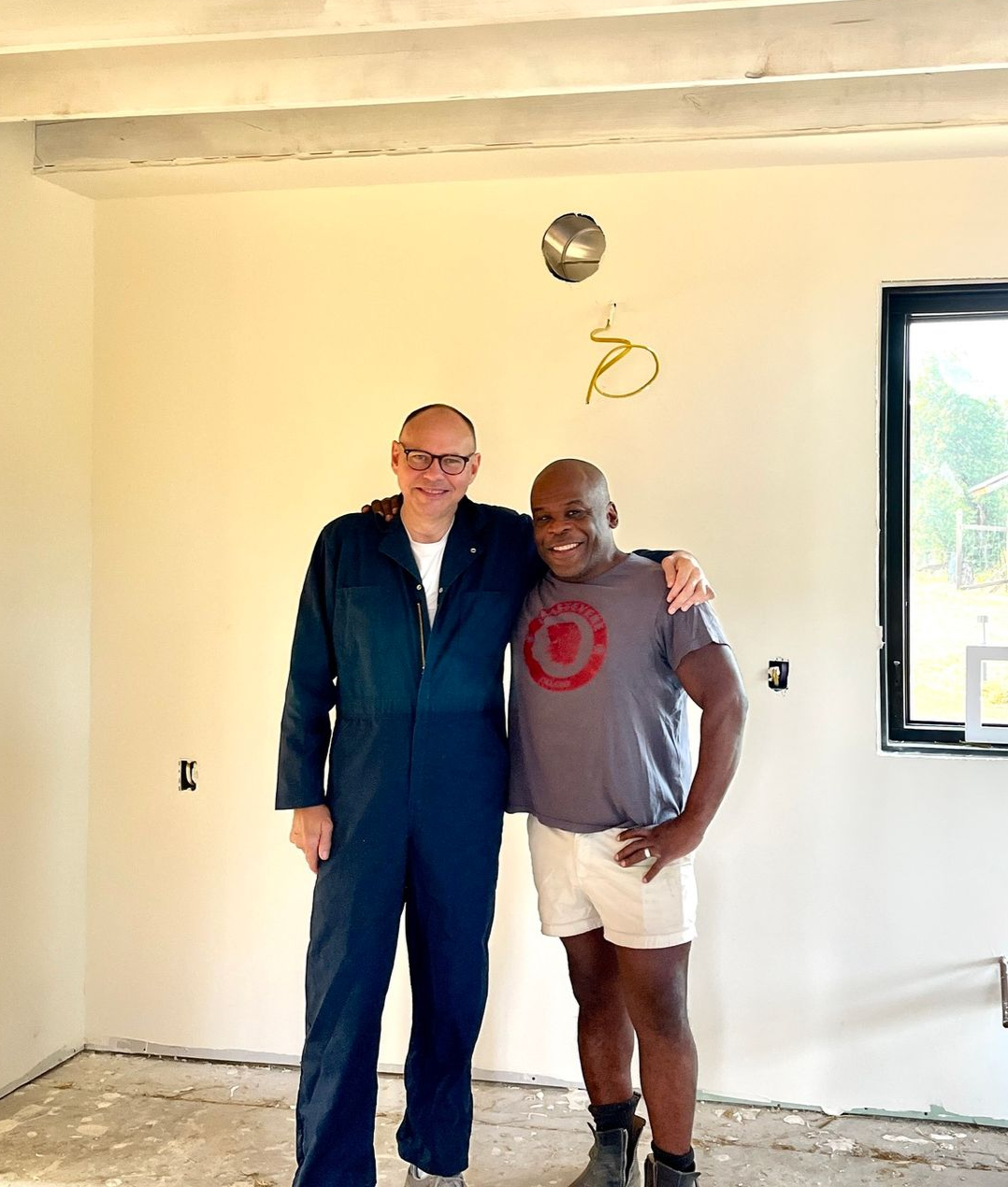 Two people standing arm-in-arm in a bright, unfinished room with beige walls and exposed light fixture wiring.