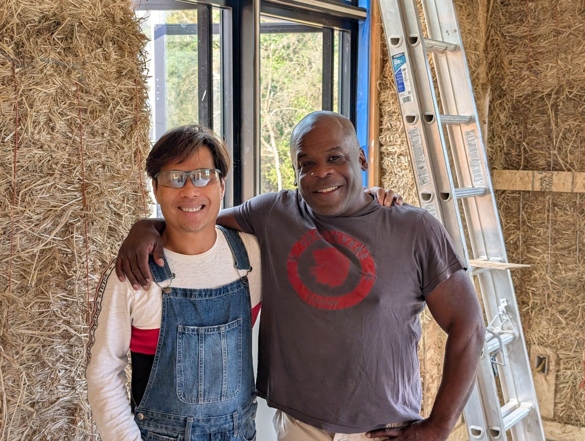 Two smiling people stand in front of straw-bale walls inside a building under construction, with a ladder to the side.