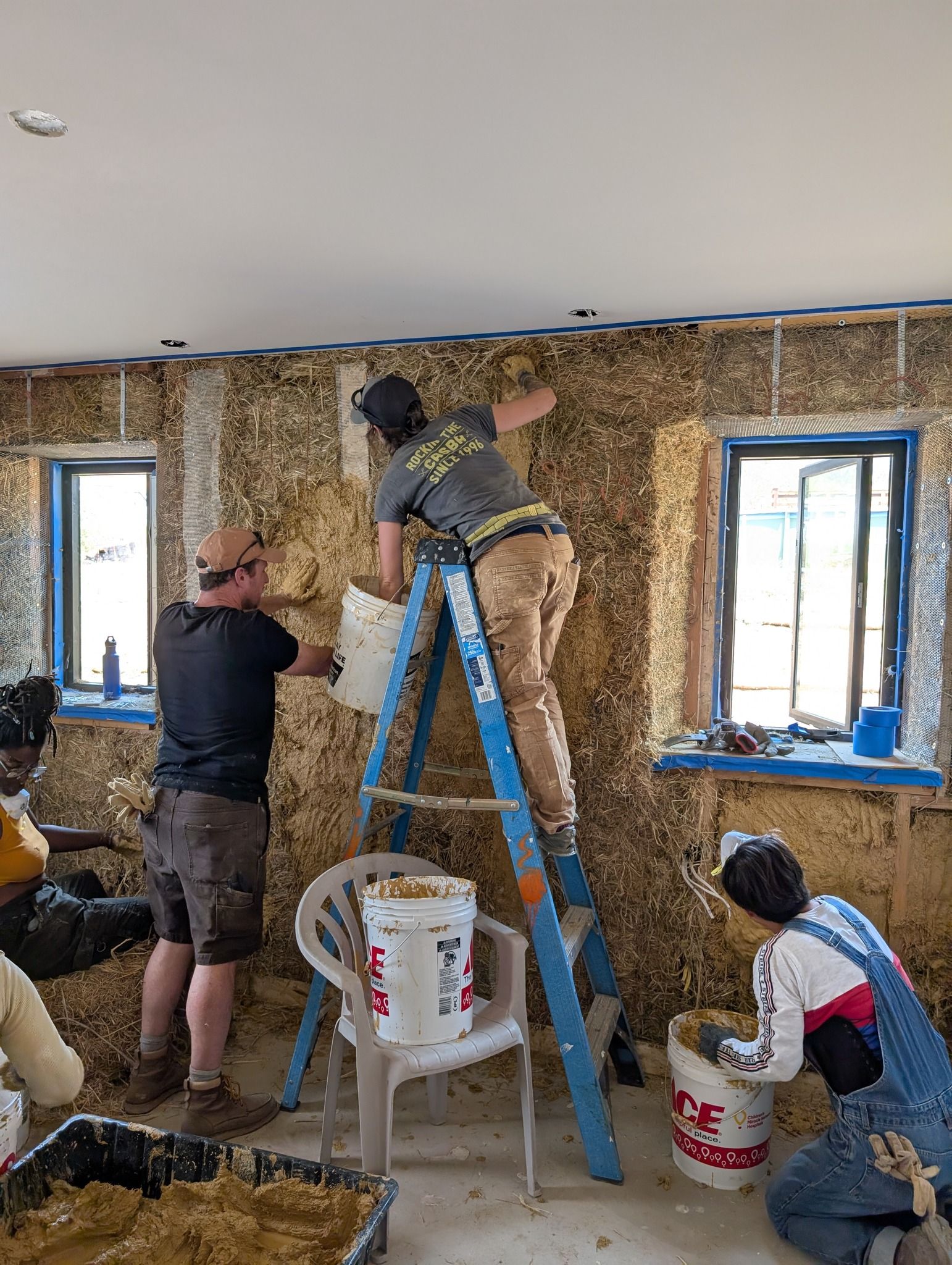 Three workers apply straw-clay insulation to interior walls in a room with two windows.