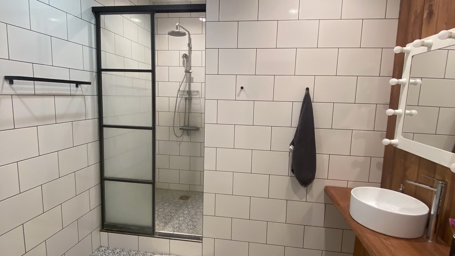 A modern bathroom with a glass shower enclosure, white brick-style tiled walls, a wooden vanity, and a round sink.