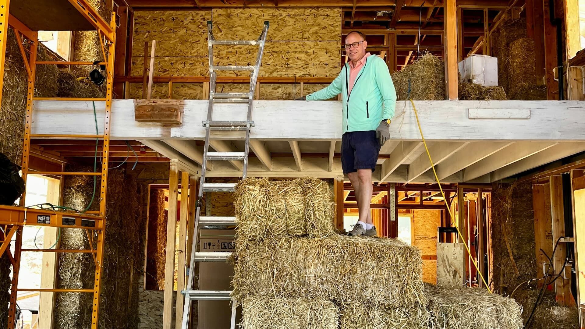 A person wearing a light blue jacket stands on a stack of hay bales inside a construction site with exposed wood framing.