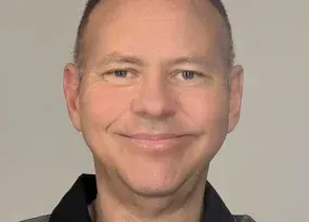 A headshot of a smiling person with short, thinning hair wearing a black collared shirt against a plain, light background.