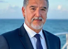 A professional headshot of a man with gray hair and a beard, wearing a suit and tie, against an ocean background.
