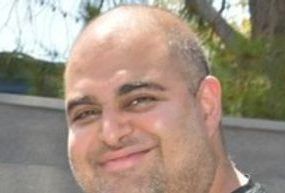 Close-up portrait of a person with a shaved head and light stubble smiling gently at the camera outdoors.