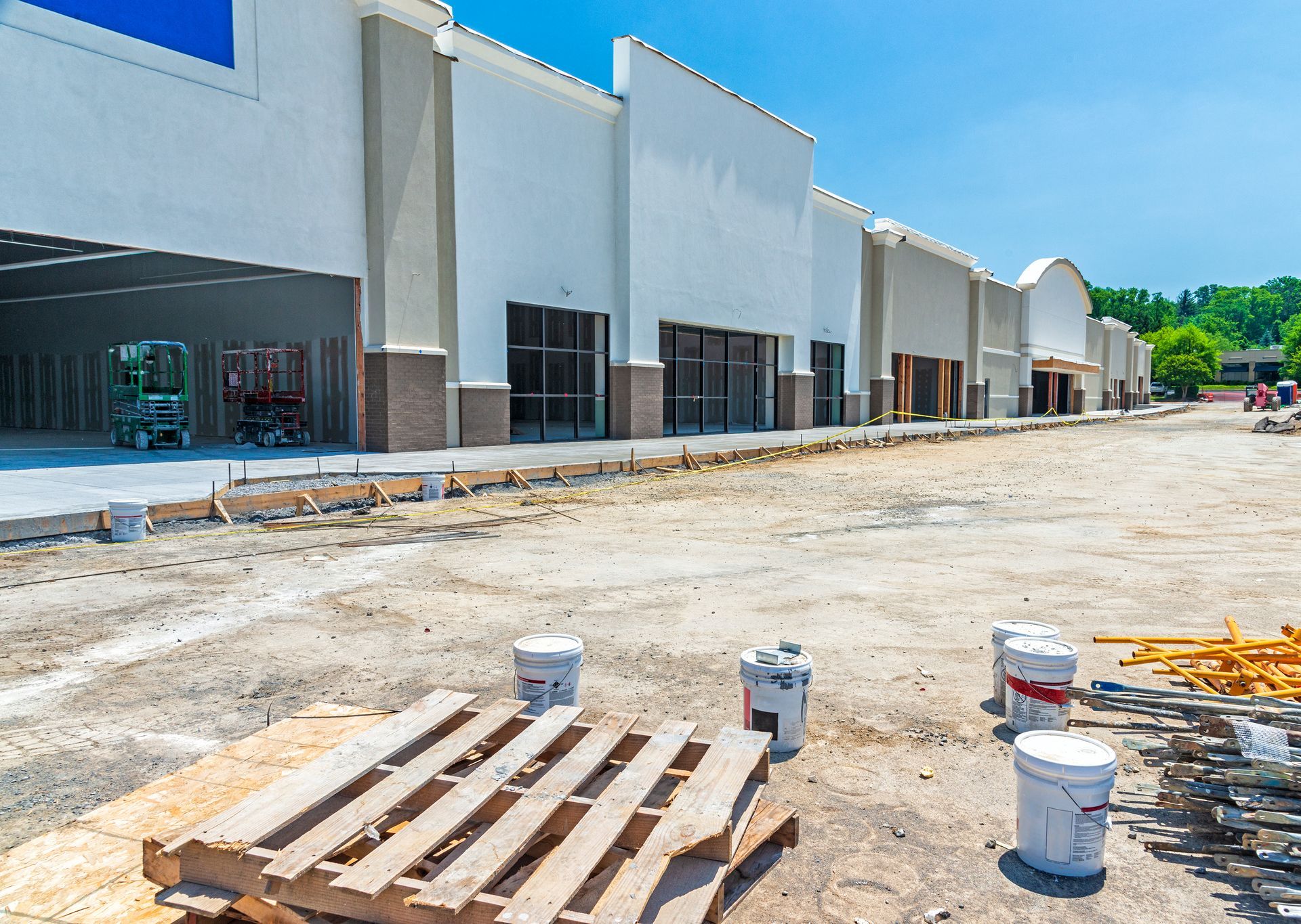 A construction site in front of a modern, multi-unit strip mall building with white walls under a bright blue sky.