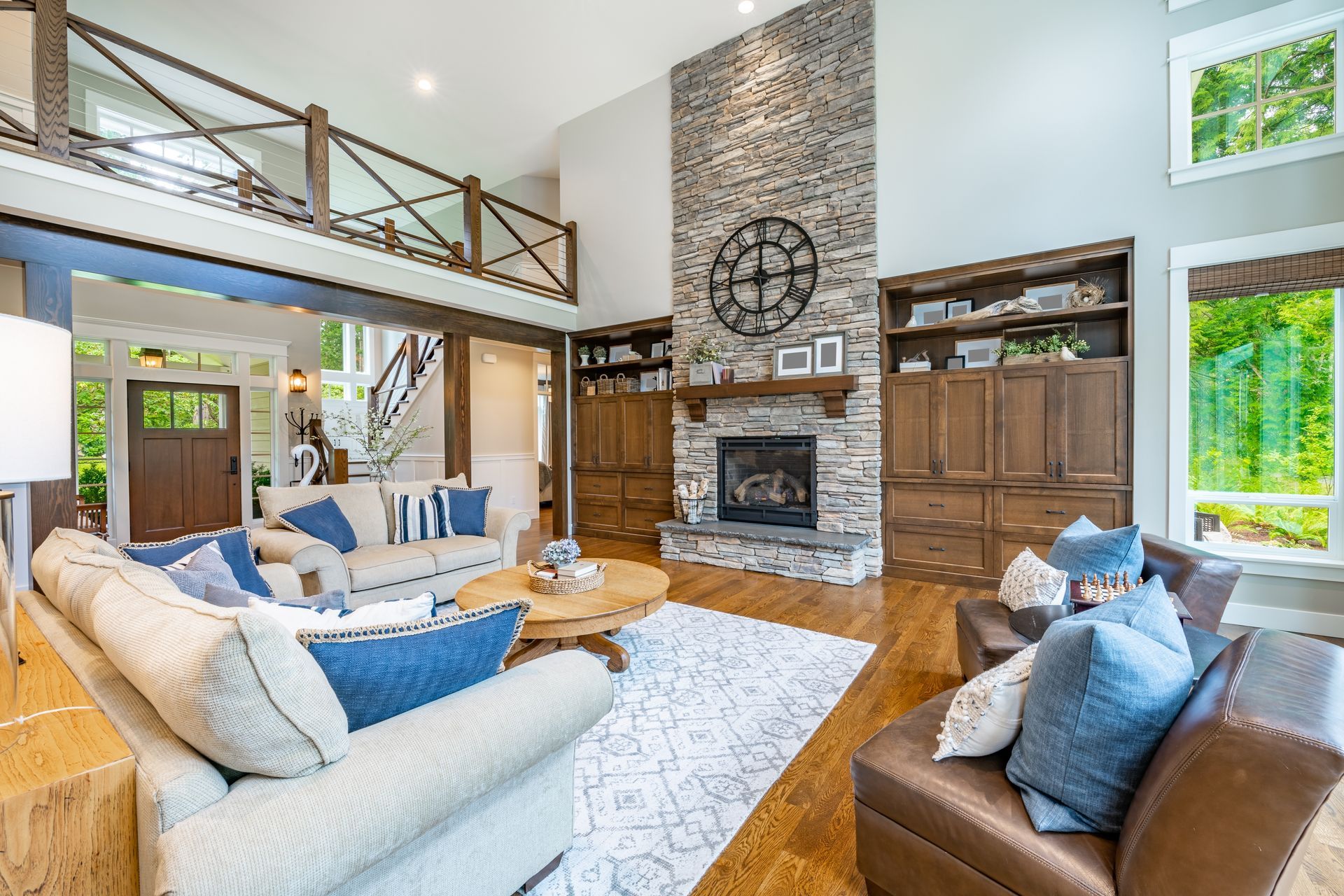 A cozy, sunlit living room with a stone fireplace, cream-colored sofas, wood trim, and a second-floor balcony.