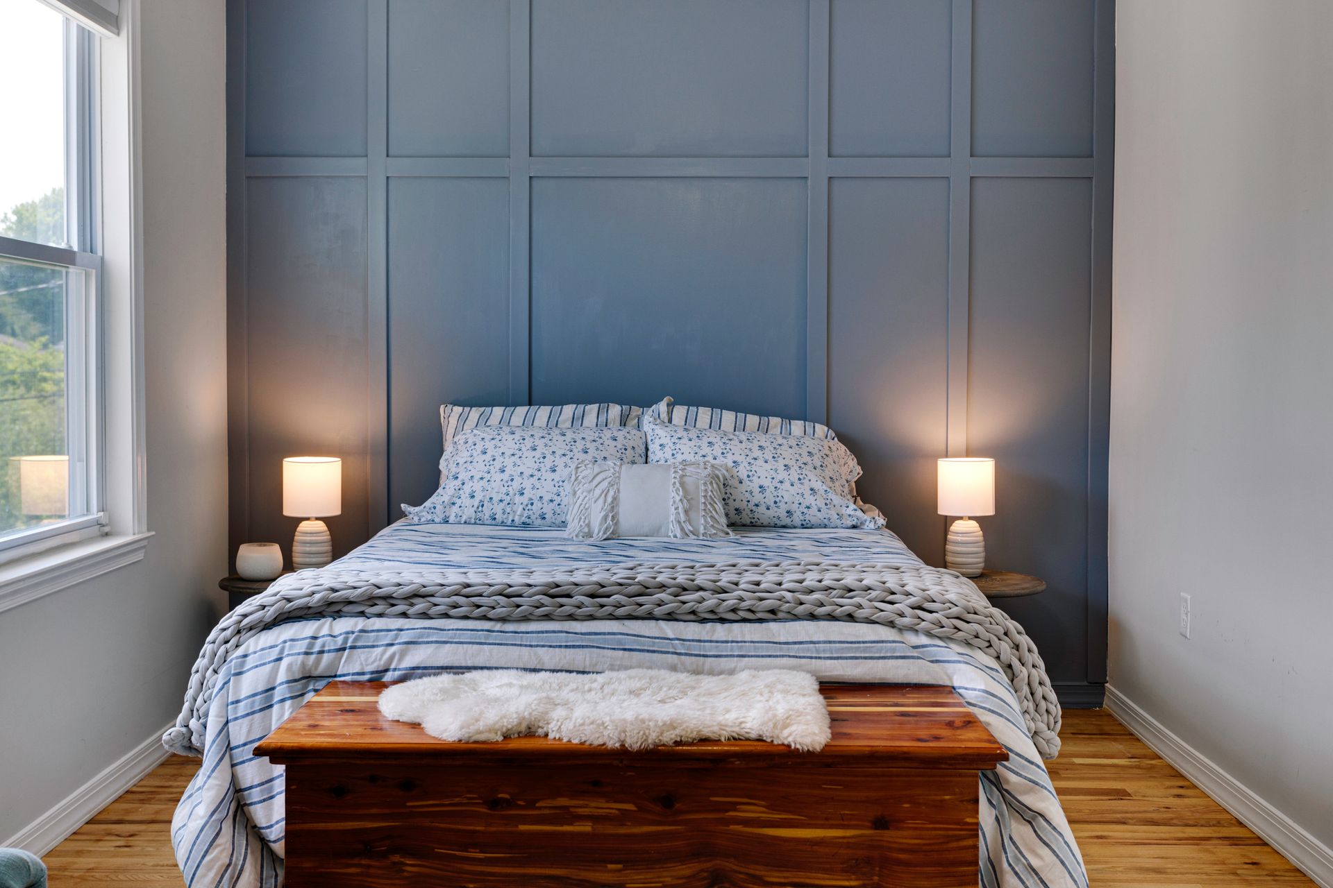 A cozy bedroom with a blue paneled accent wall, a bed with patterned bedding, two small lamps, and a wooden chest.