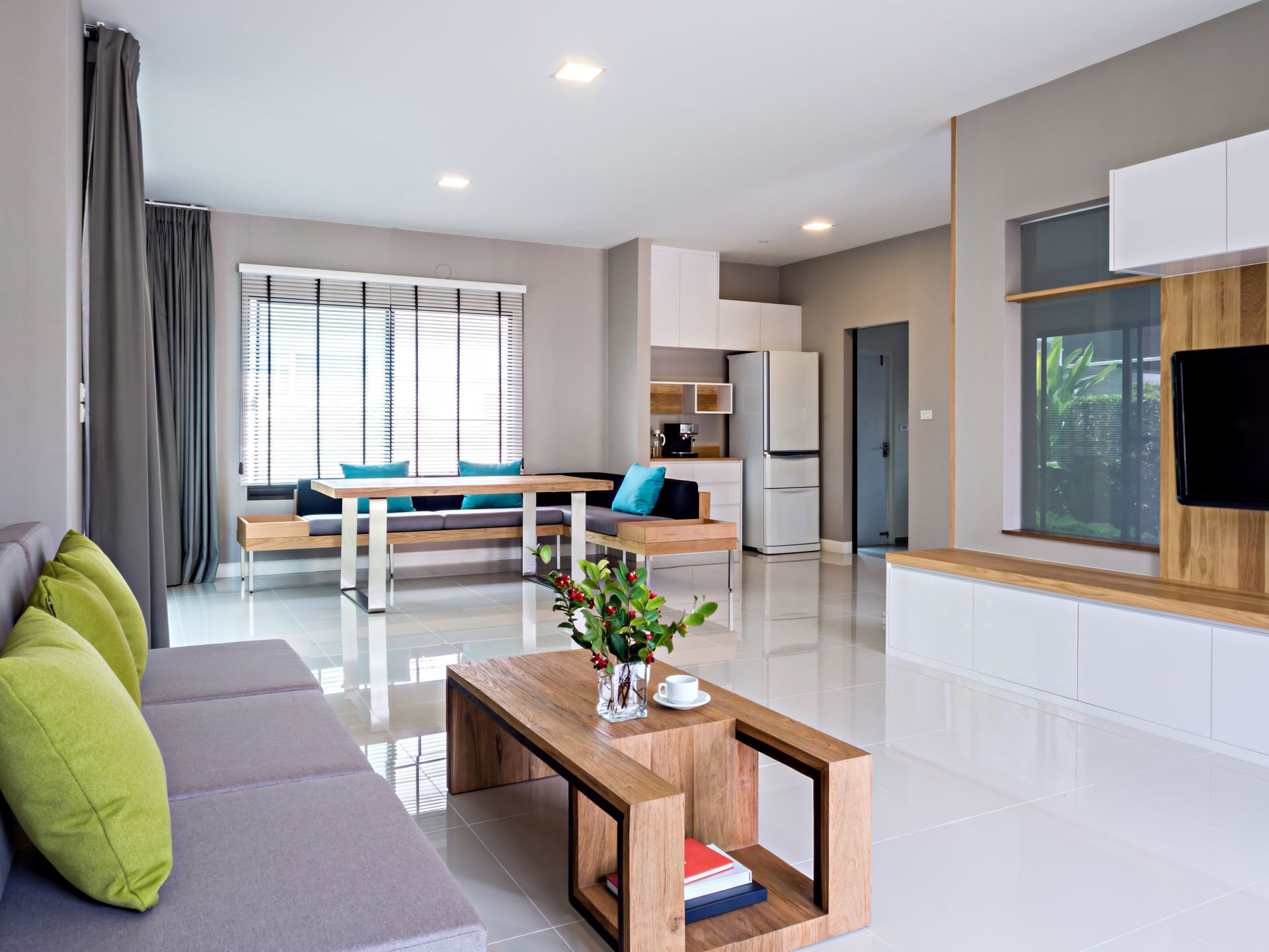 A modern, minimalist living room with a gray sofa, green pillows, a wooden coffee table, and a dining area in the back.