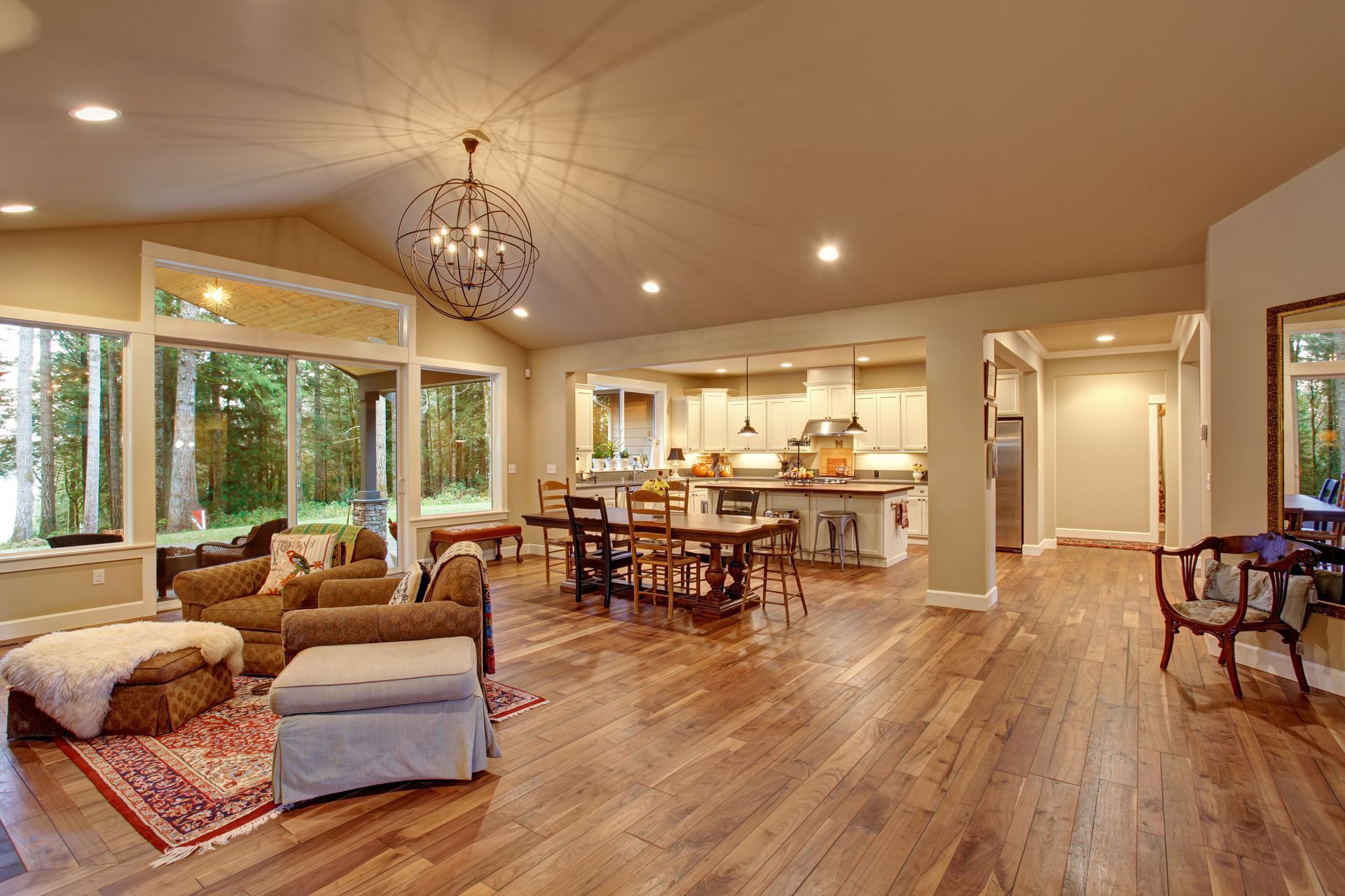 An open-plan living and dining area with hardwood floors, a stone fireplace, large windows, and a rustic chandelier.