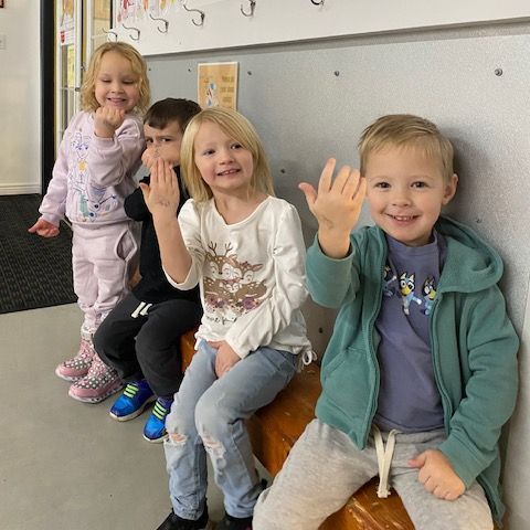 A group of children are sitting on a bench and waving at the camera.