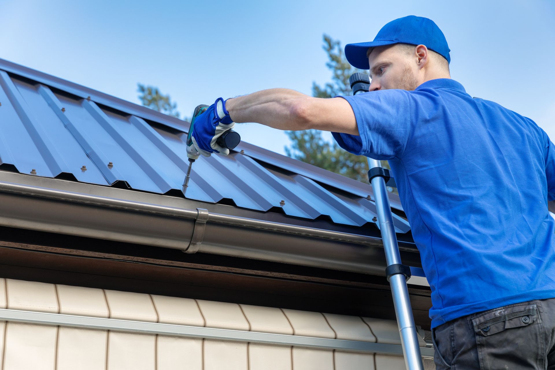 Roofer with blue uniform and cap drills metal tile on roof, with blue sky background.