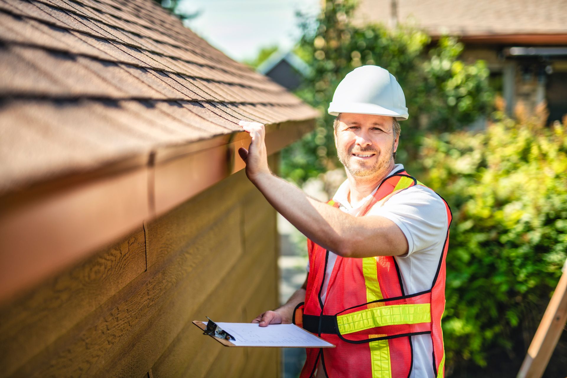 Man in hard hat and safety vest inspecting a roof gutter, holding clipboard.