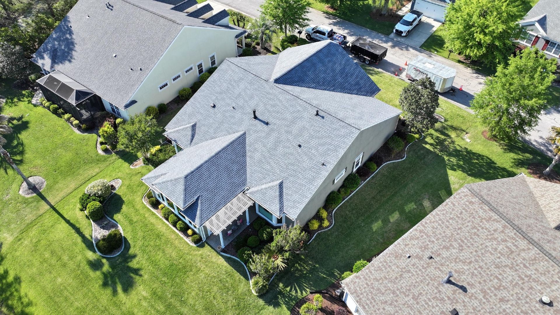 Aerial view of a white multi-roof house with gray shingles and metal accents, surrounded by a yard with green landscaping.