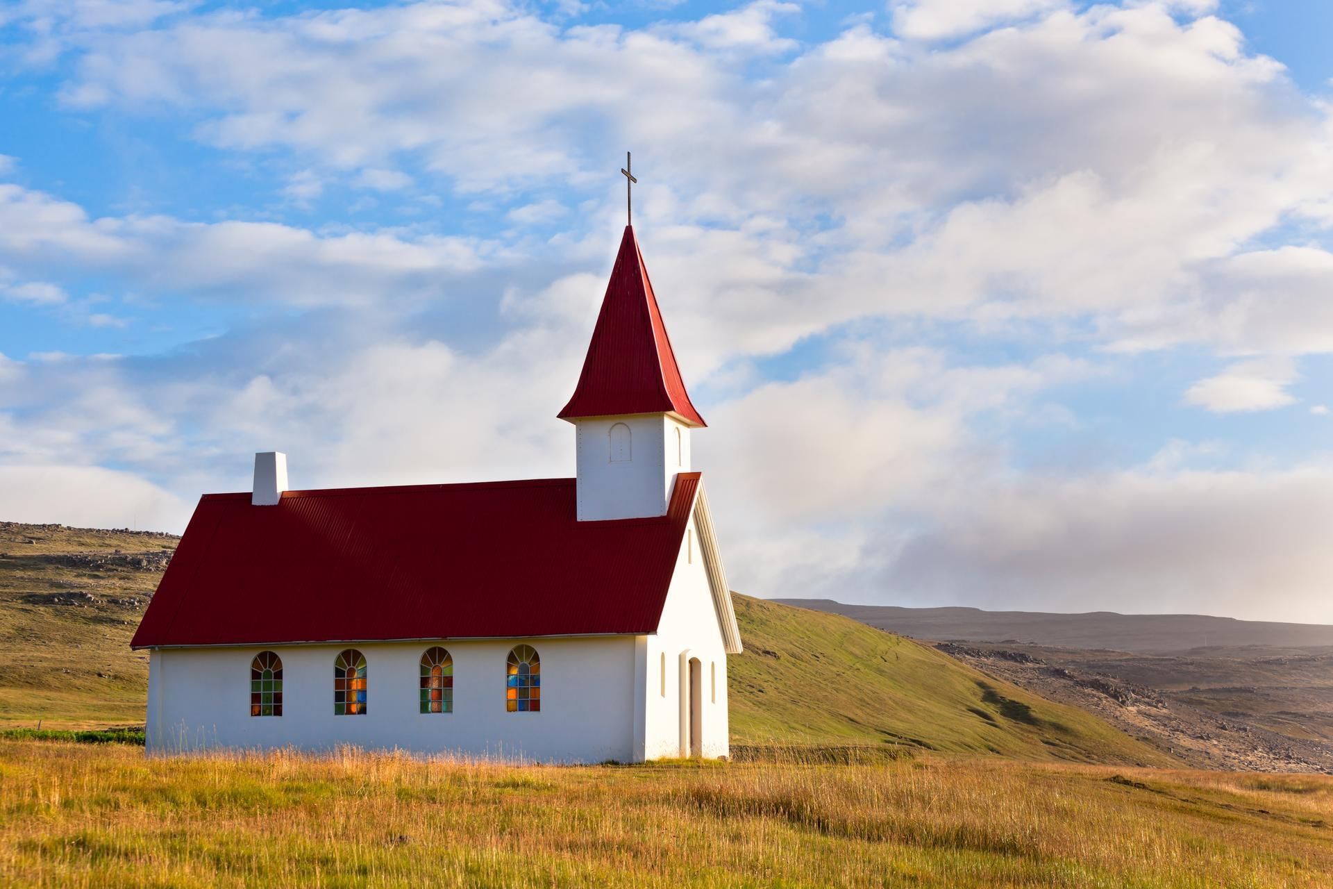 A small white church with a red roof is sitting on top of a grassy hill.