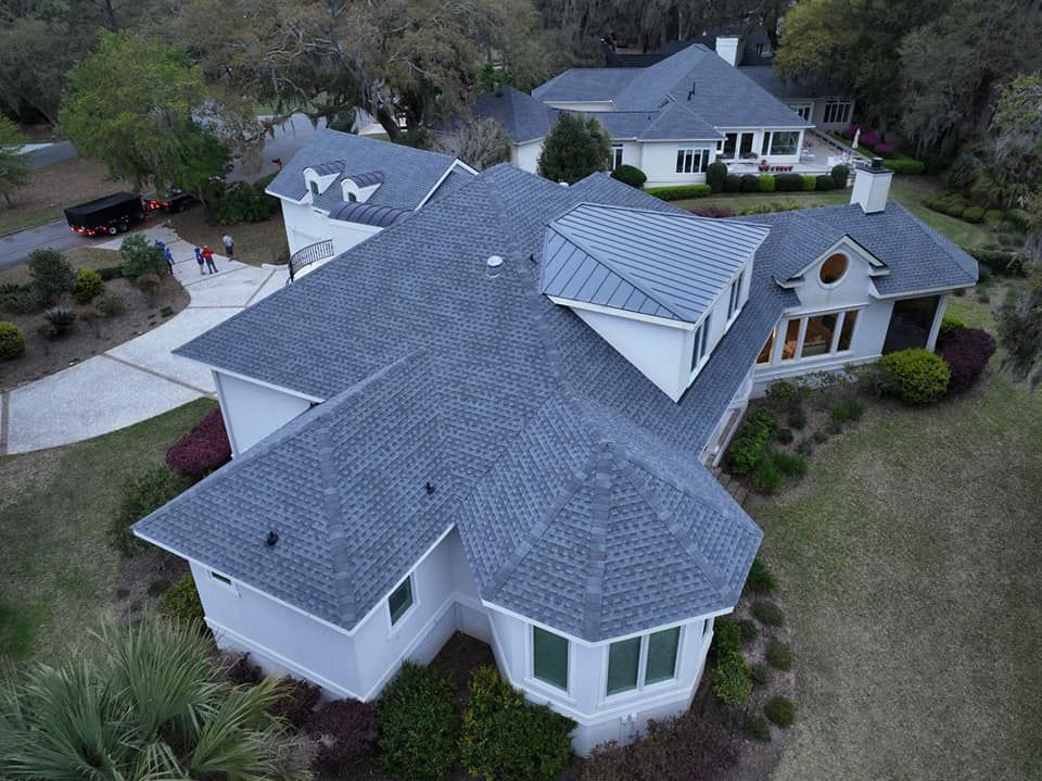 Aerial view of a white multi-roof house with gray shingles and metal accents, surrounded by a yard with green landscaping. Aerial view of a white multi-roof house with gray shingles and metal accents, surrounded by a yard with green landscaping.
