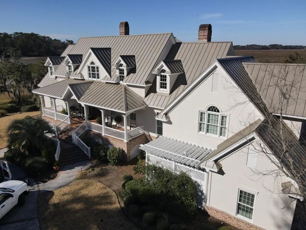 An aerial view of a large, cream-colored estate house with a textured, patterned metal roof, chimneys, and landscaping.