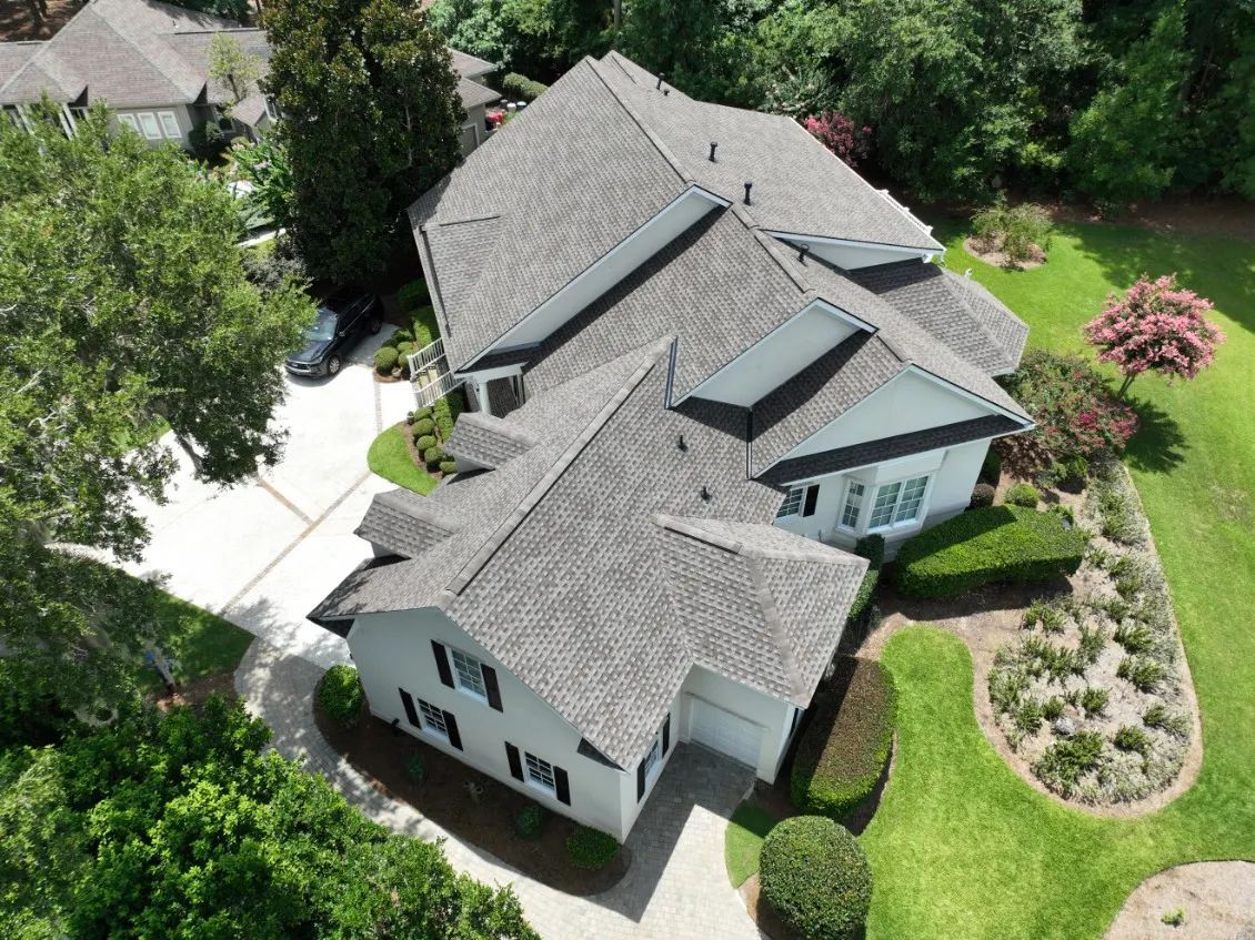 An aerial view of a white, multi-level house with a gray shingled roof, surrounded by green lawns and trees. An aerial view of a white, multi-level house with a gray shingled roof, surrounded by green lawns and trees.