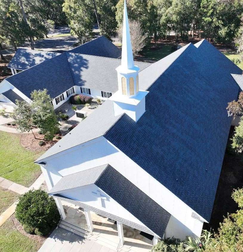 An aerial view of a white church with a dark gray shingled roof, steeple, and surrounding trees and walkways.