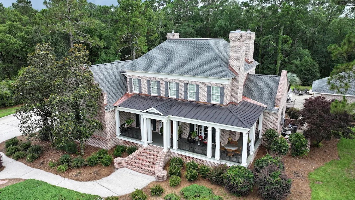 An elevated view of a brick house with a dark metal porch roof and a shingled roof, surrounded by trees and landscaping.
