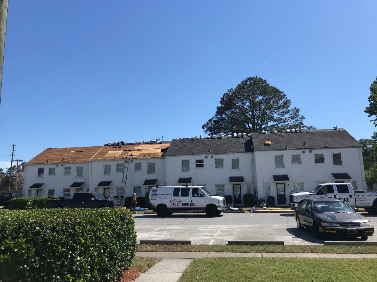 Row of white apartment buildings, roofers on the roof, work vans parked below, sunny day.