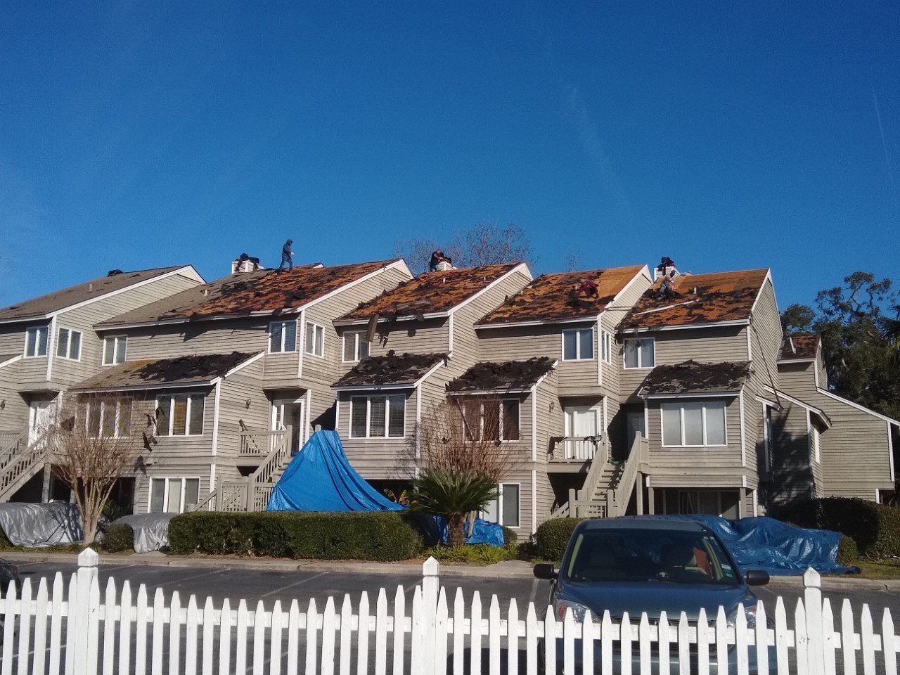 Row of townhouses with roofing being replaced; blue tarps on ground. Clear blue sky overhead.