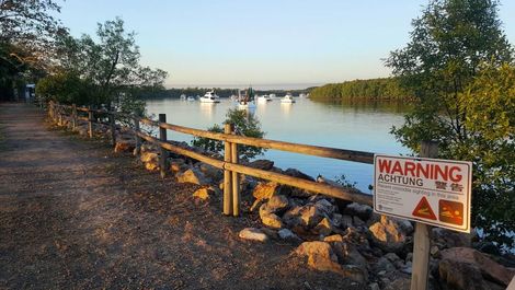 A Warning Sign Is Sitting Next To A Wooden Fence Next To A Body Of Water — Mission Solar And Electrical In Cardwell, QLD