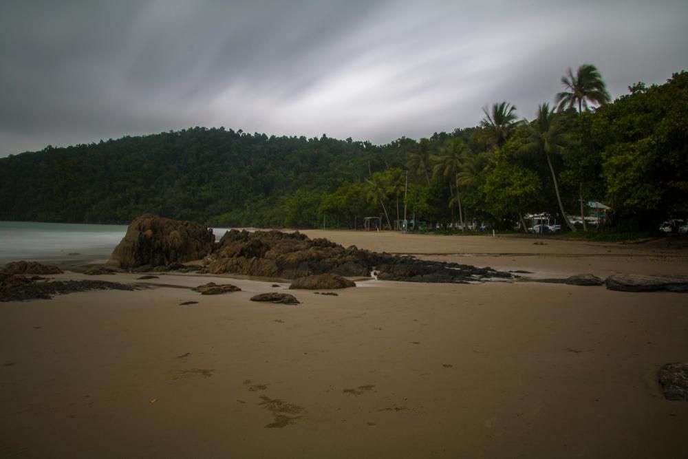 A Beach With Palm Trees And Mountains In The Background On A Cloudy Day  — Mission Solar And Electrical In Innisfail, QLD