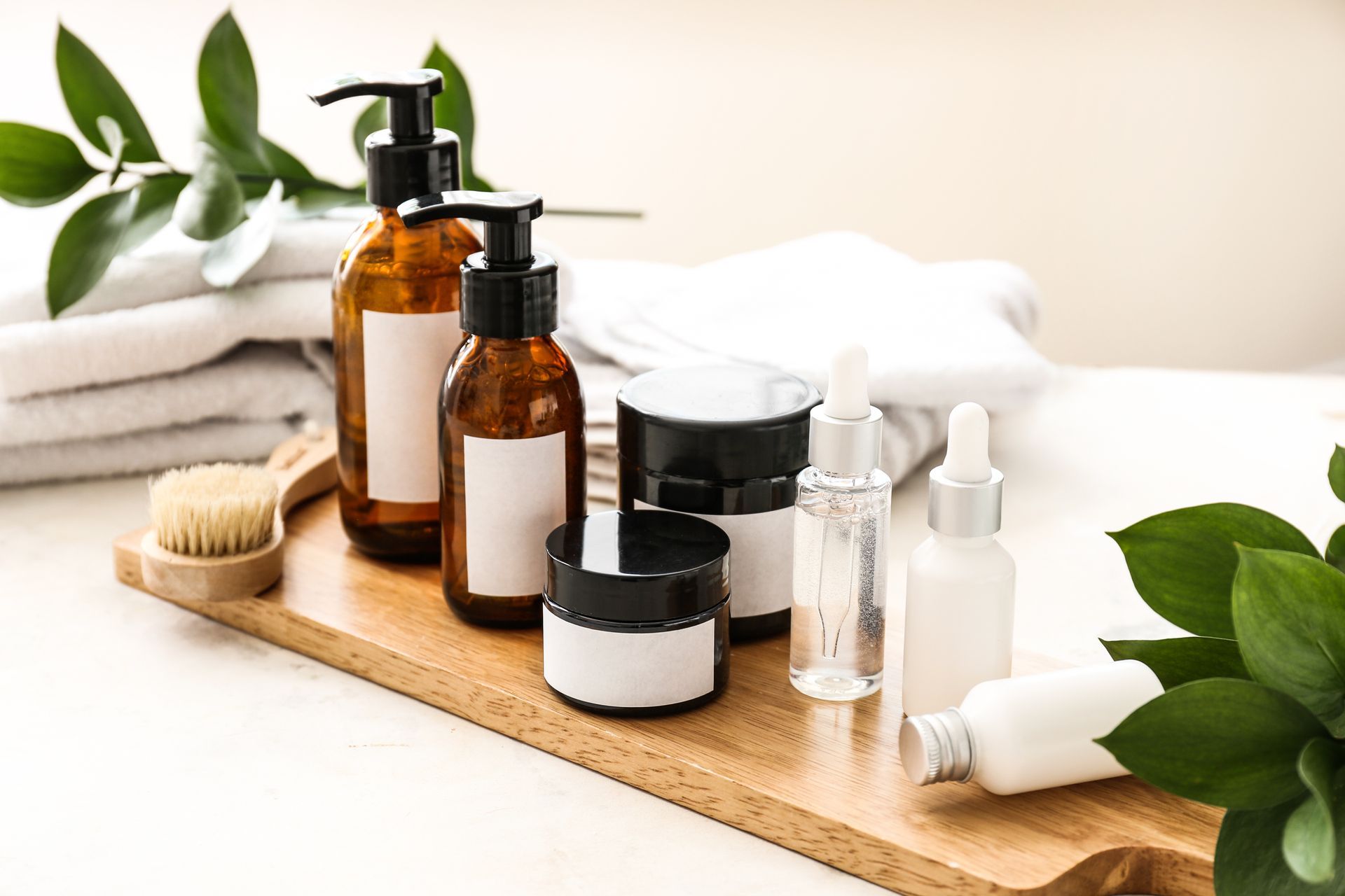 Skincare products on a wooden tray with towels and greenery in a bright setting.