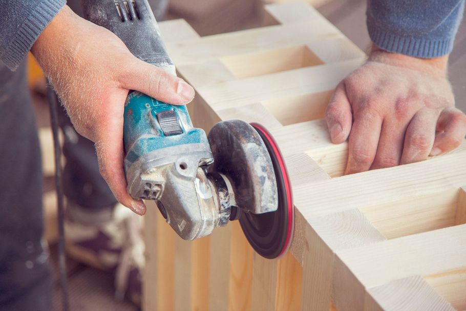 Hands using an angle grinder to sand a wooden board in a workshop.