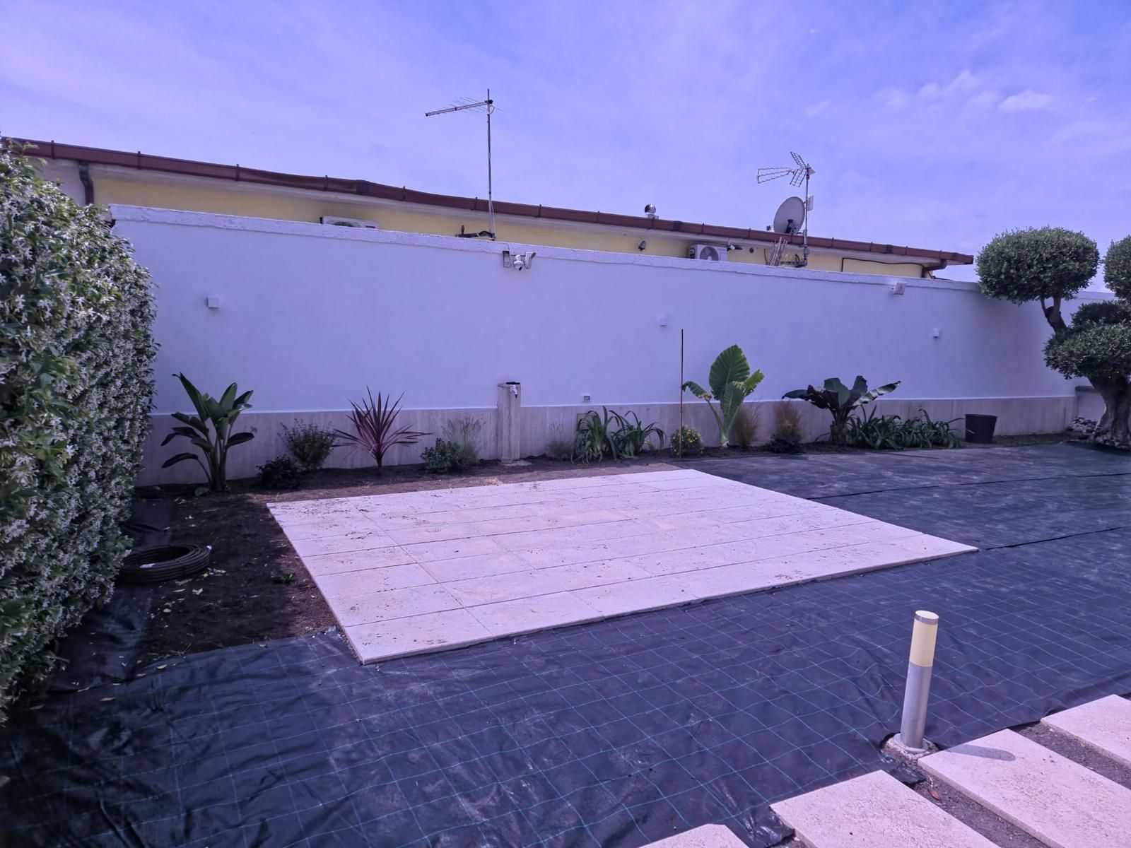 Cortile con area pavimentata rettangolare, piante e un muro bianco sotto un cielo azzurro.