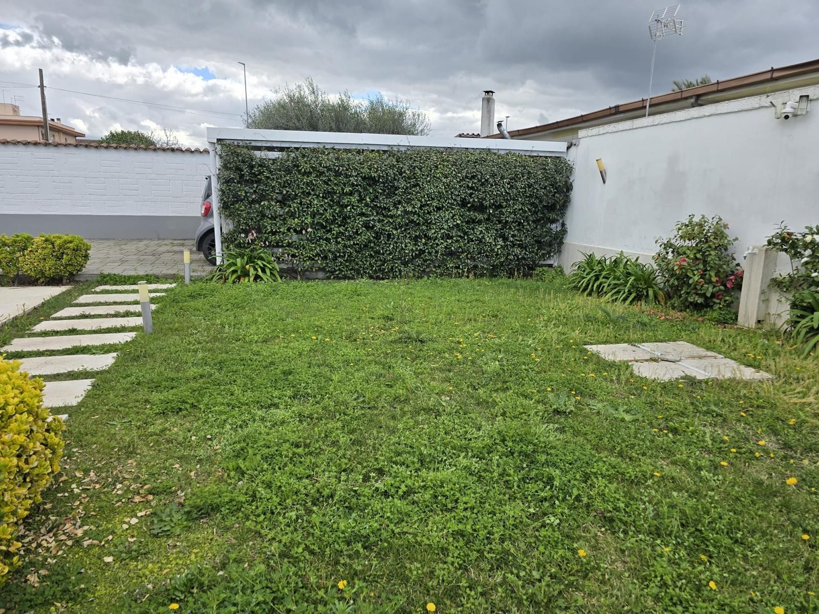 Prato davanti a una siepe e muri bianchi sotto un cielo nuvoloso. Sentiero in pietra sulla sinistra.