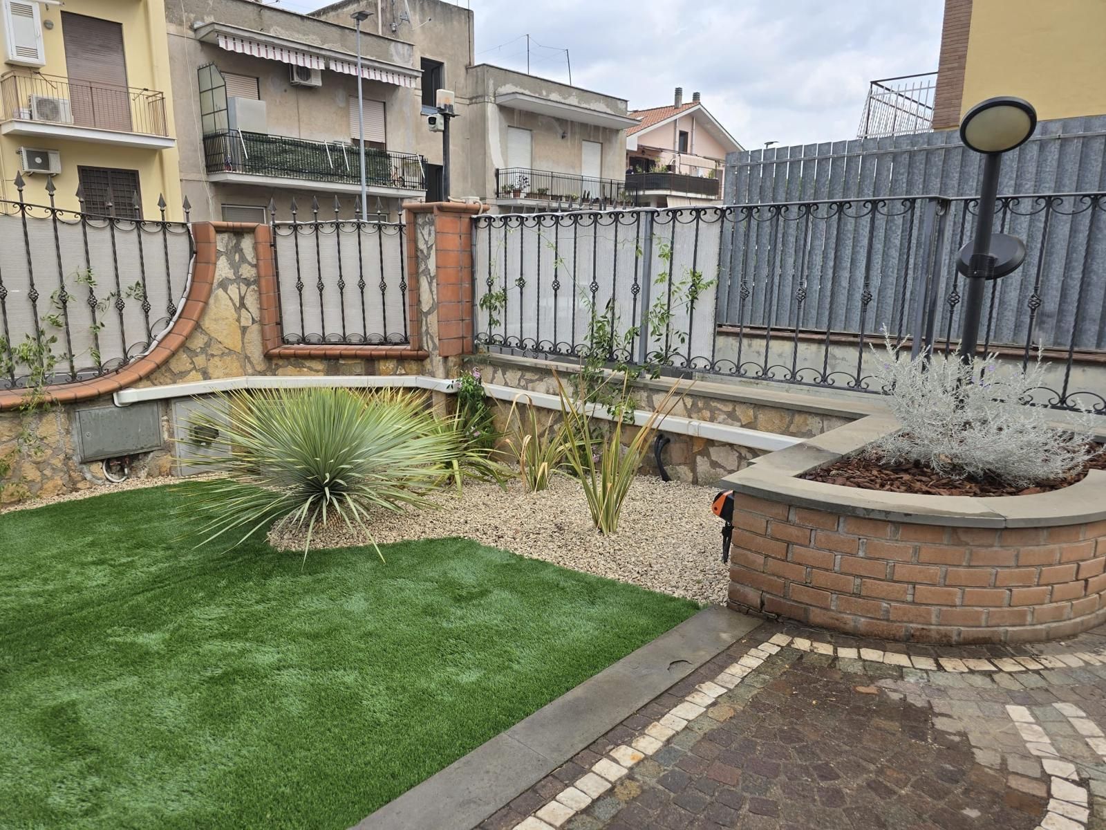 Un piccolo cortile con erba sintetica, ghiaia e mattoni, circondato da una recinzione metallica.