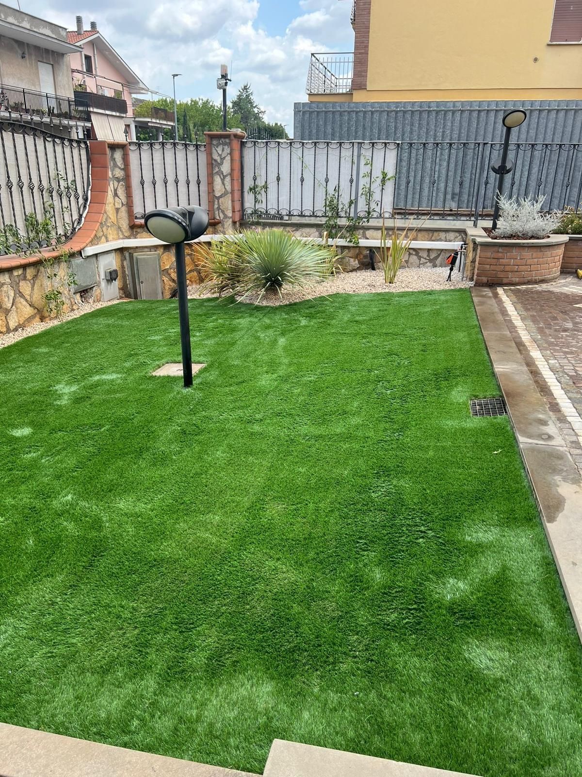 Prato artificiale verde in un cortile con piccoli arbusti, bordure in mattoni e recinzioni.