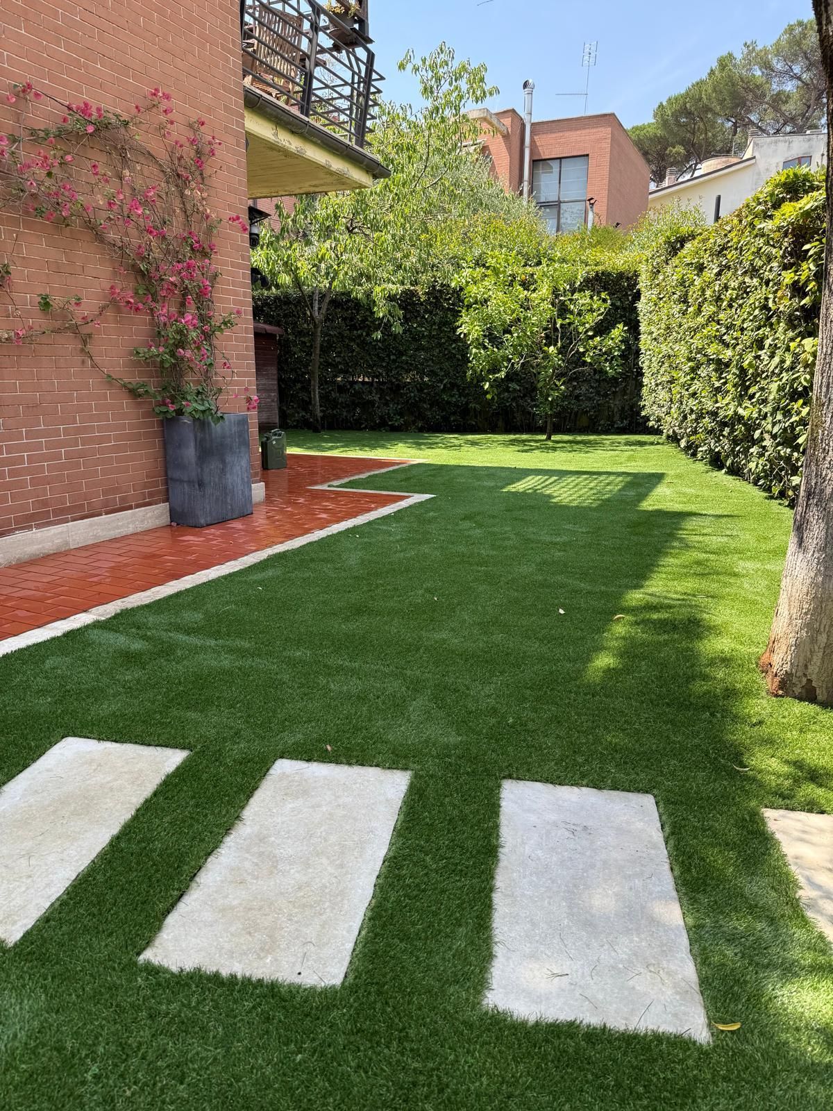 Prato verde con gradini in pietra, casa in mattoni rossi con vite fiorita e alte siepi verdi.