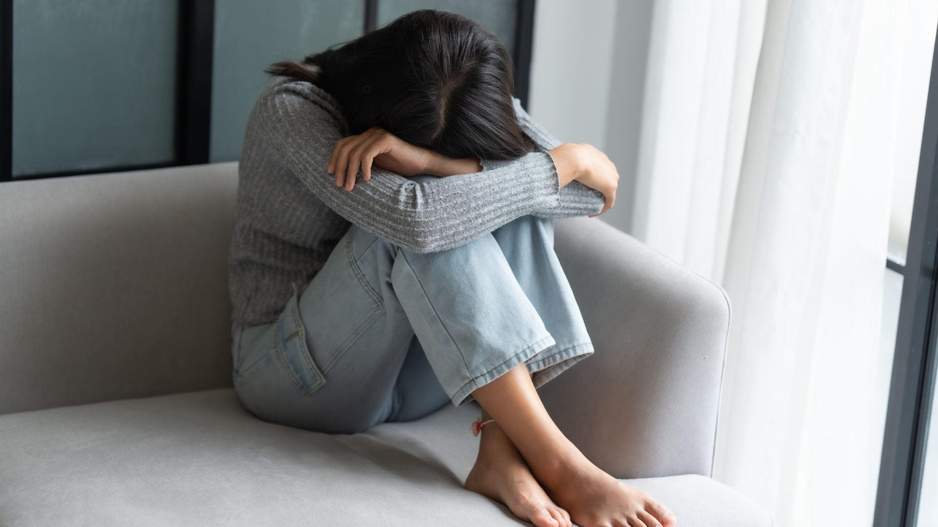 A woman sitting on a couch with her head down, holding her arms, looking overwhelmed by emotions, reflecting symptoms of depression.